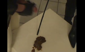 Blonde Woman Feeds Man Mask with Shit on Bed
